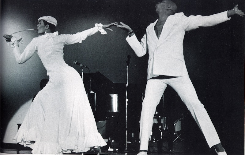Carmen de Lavallade in a long white dress smokes a long pipe. Her outstretched hand is held by her husband Geoffrey Hold also dressed in white
